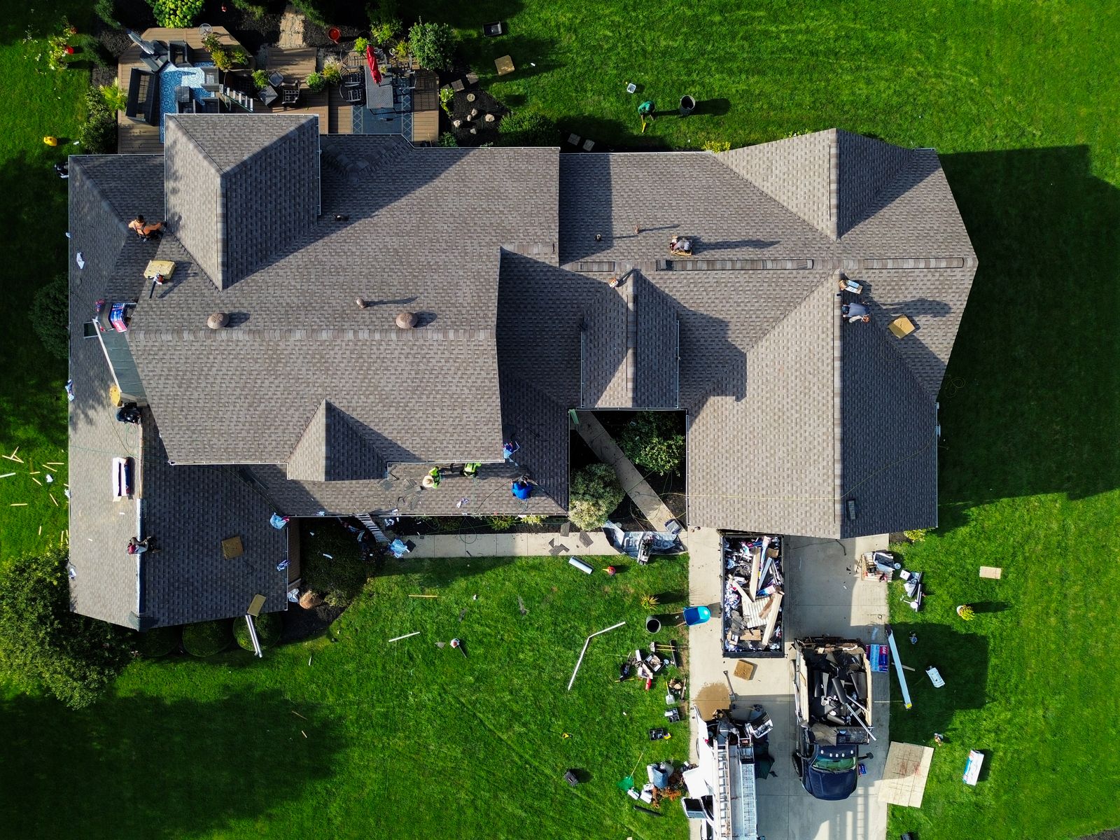Overhead of a roof replacement in progress with crew across multiple planes and a loaded dumpster in the driveway — Hardin County, Ohio